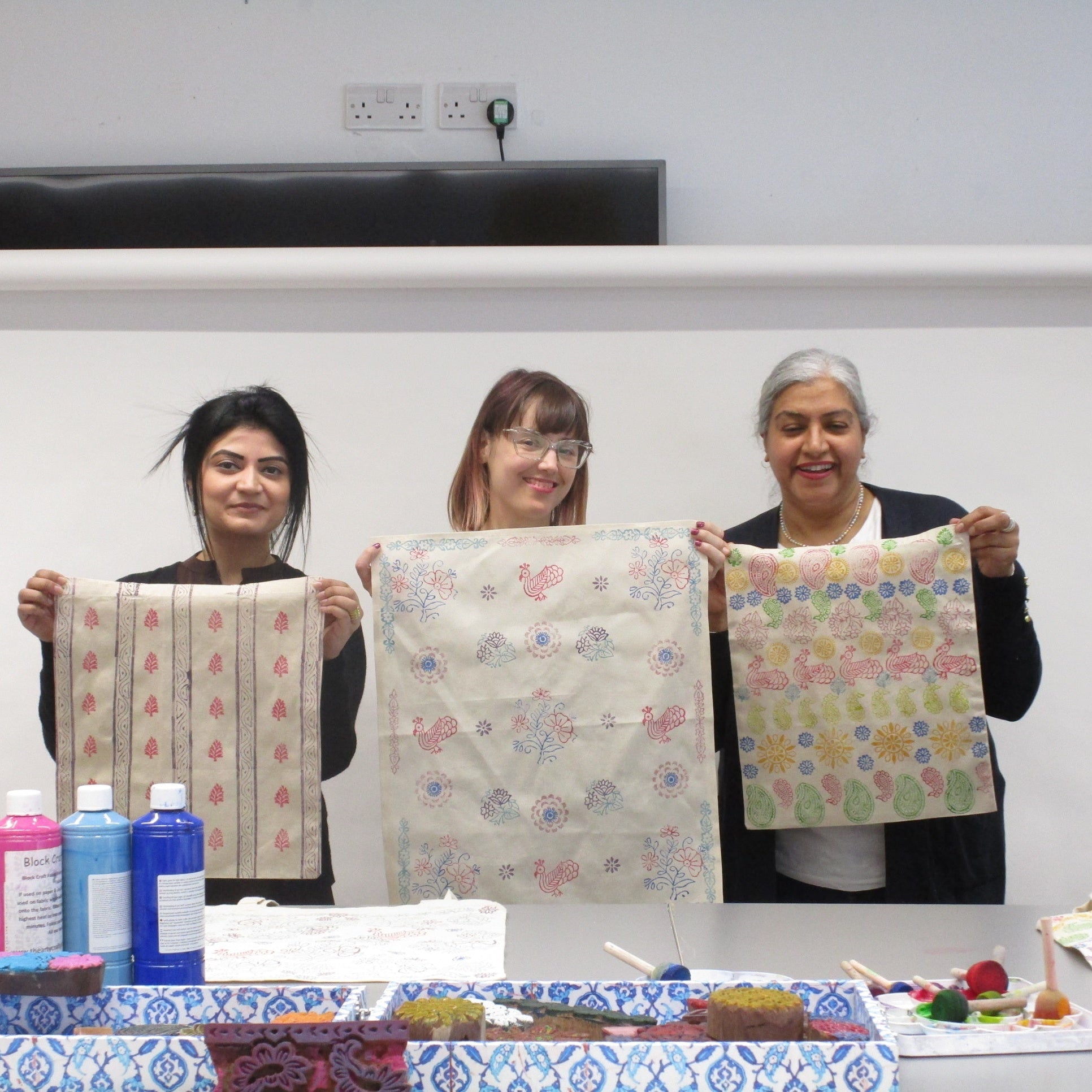 Three women holding up fabric with floral patterns in a classroom setting.