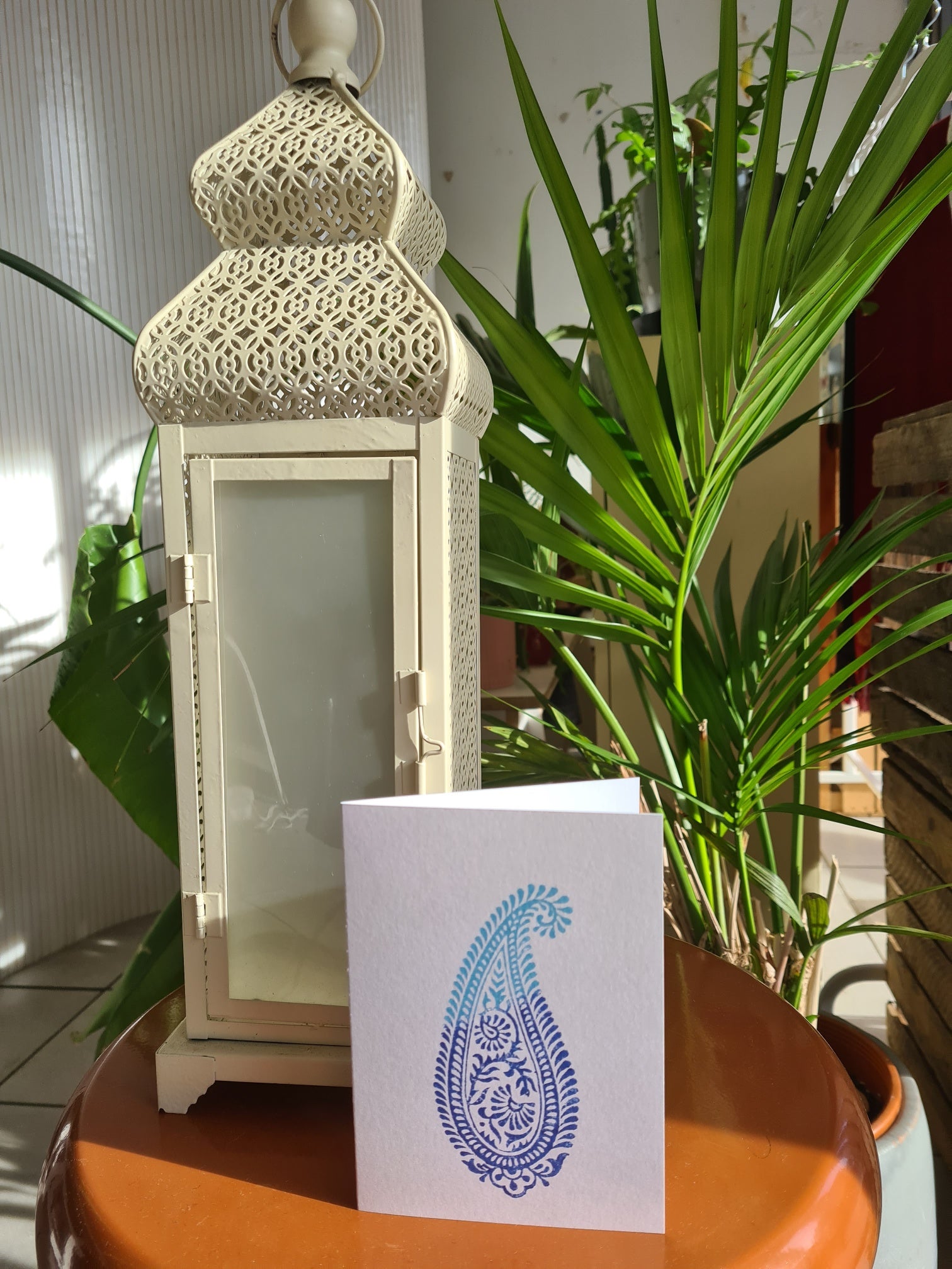 Decorative lantern and card with paisley design on a table with plants in the background