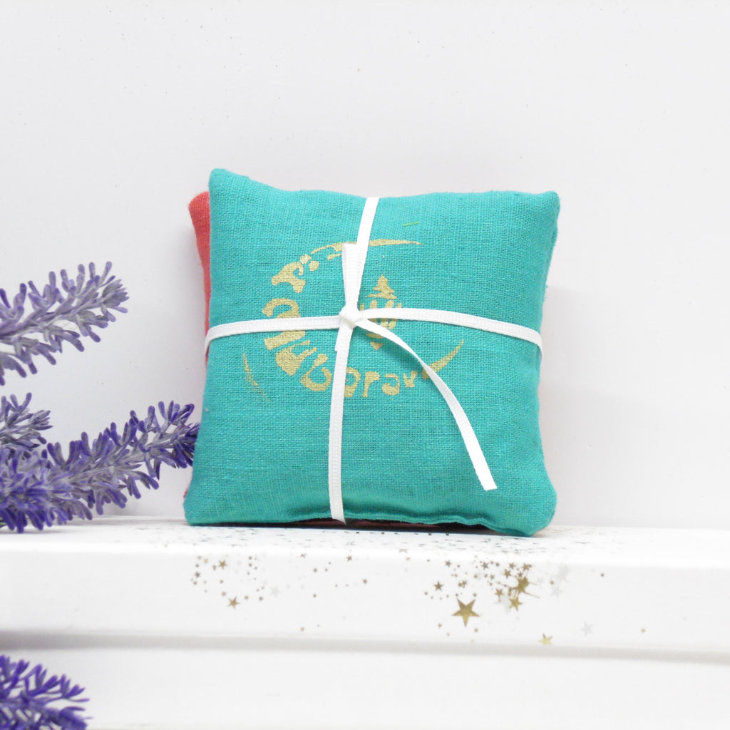 Turquoise pillow with white ribbon and gold text on a white surface with lavender flowers.
