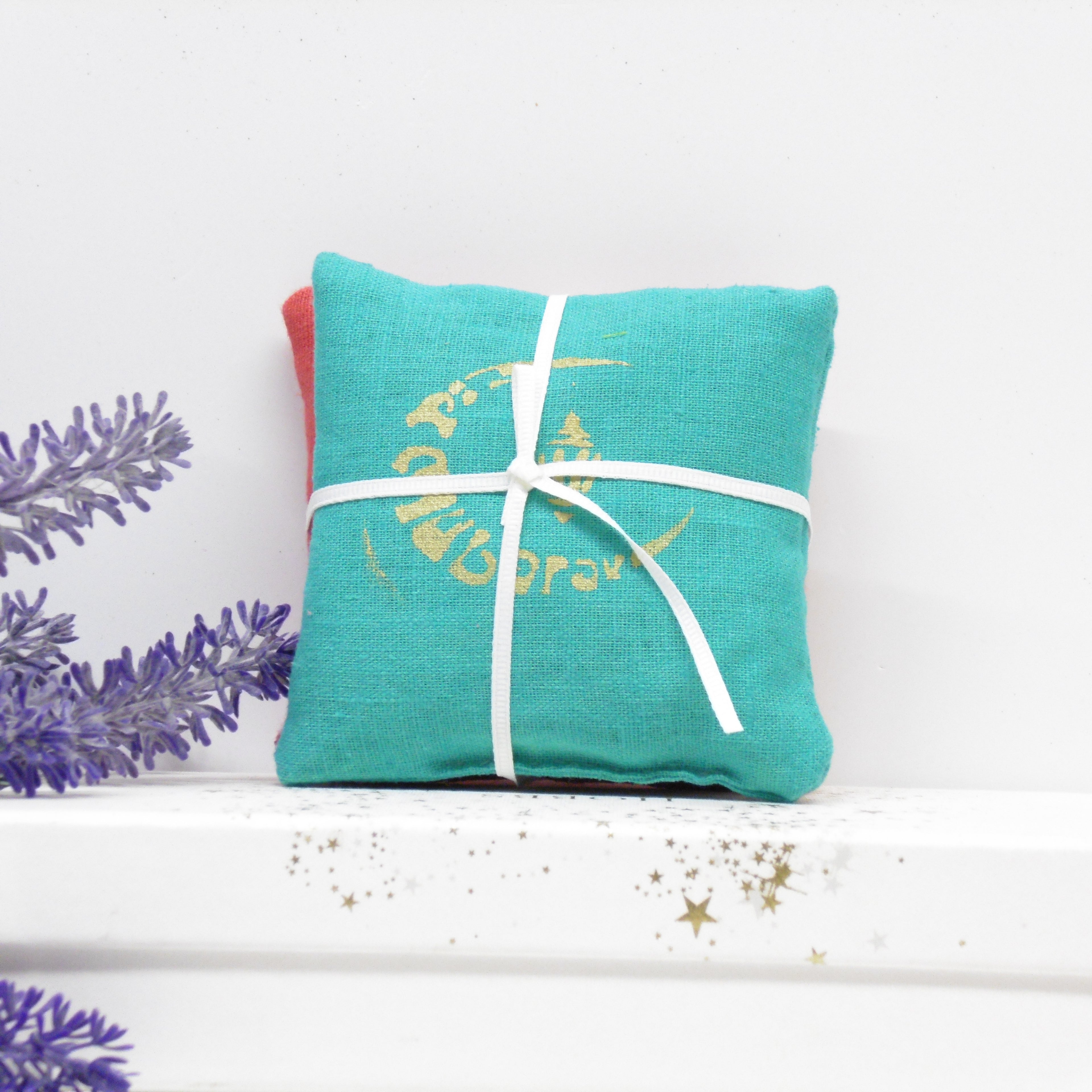 Turquoise pillow with white ribbon and gold text on a white surface with lavender flowers.