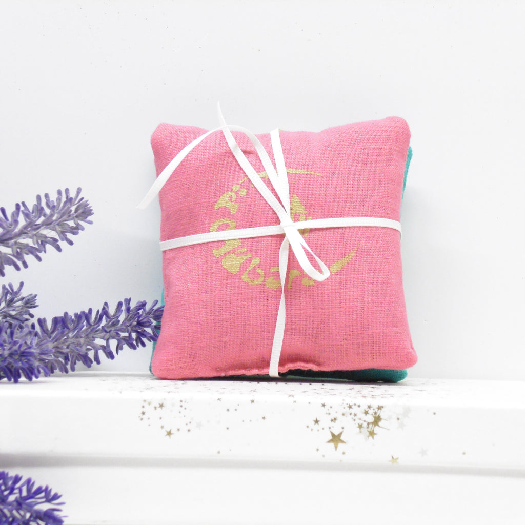 Pink pillow with white ribbon and gold text on a white surface with lavender flowers.