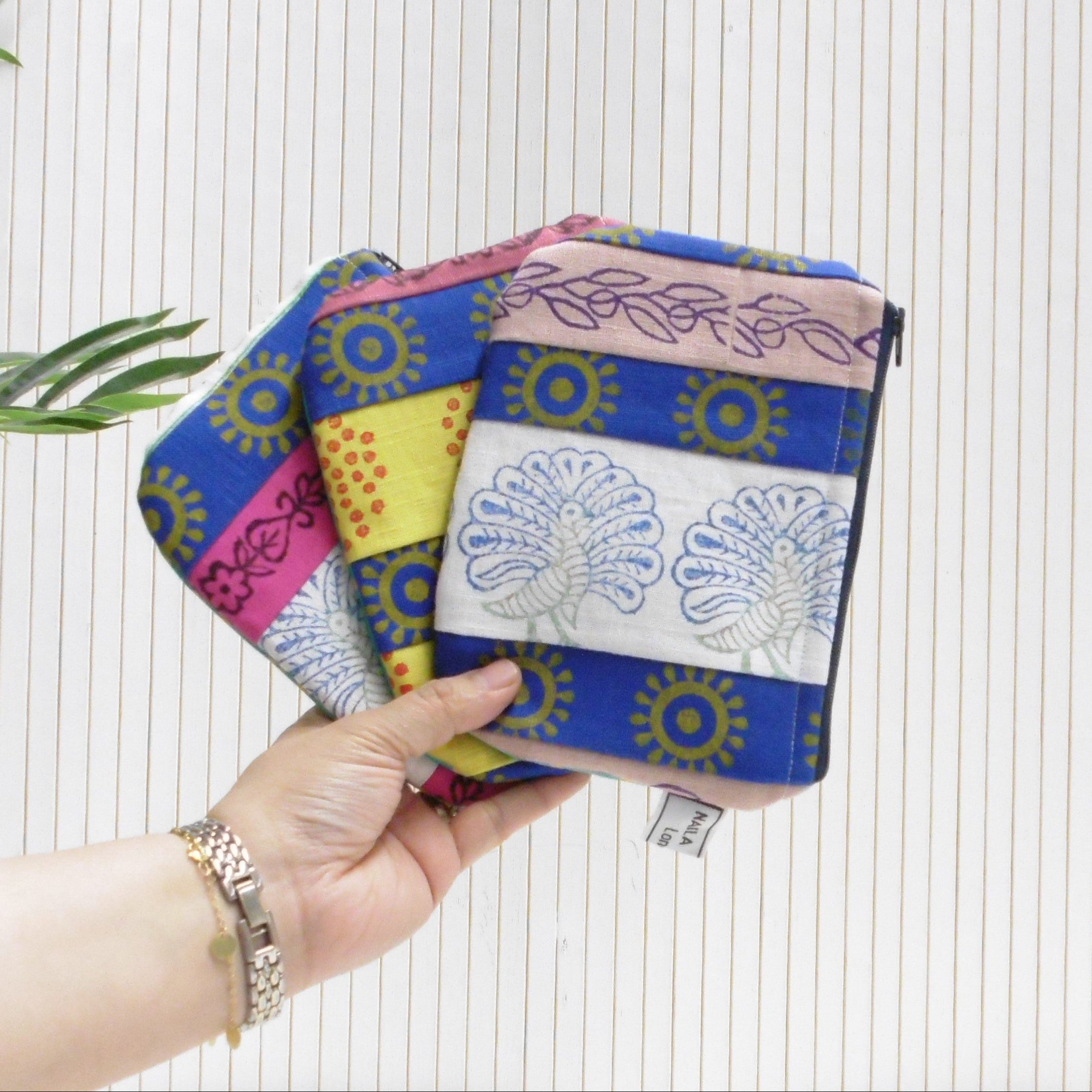 Hand holding a colorful fabric pouch against a white background with green leaves.