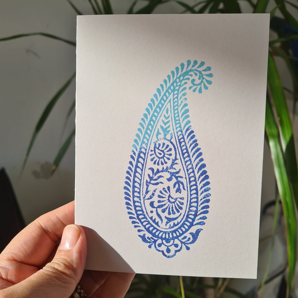Hand holding a card with a blue paisley design against a plant background