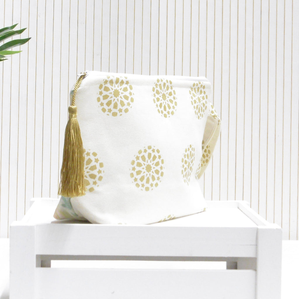 White pouch with gold floral pattern and tassel on a white surface with a light wooden background