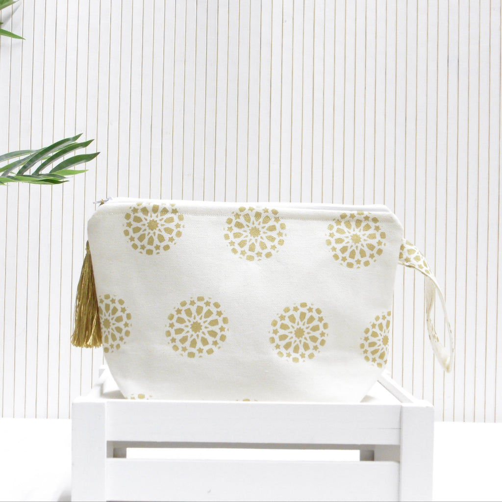 White pouch with gold floral patterns and a tassel on a white chair against a light wooden panel background.